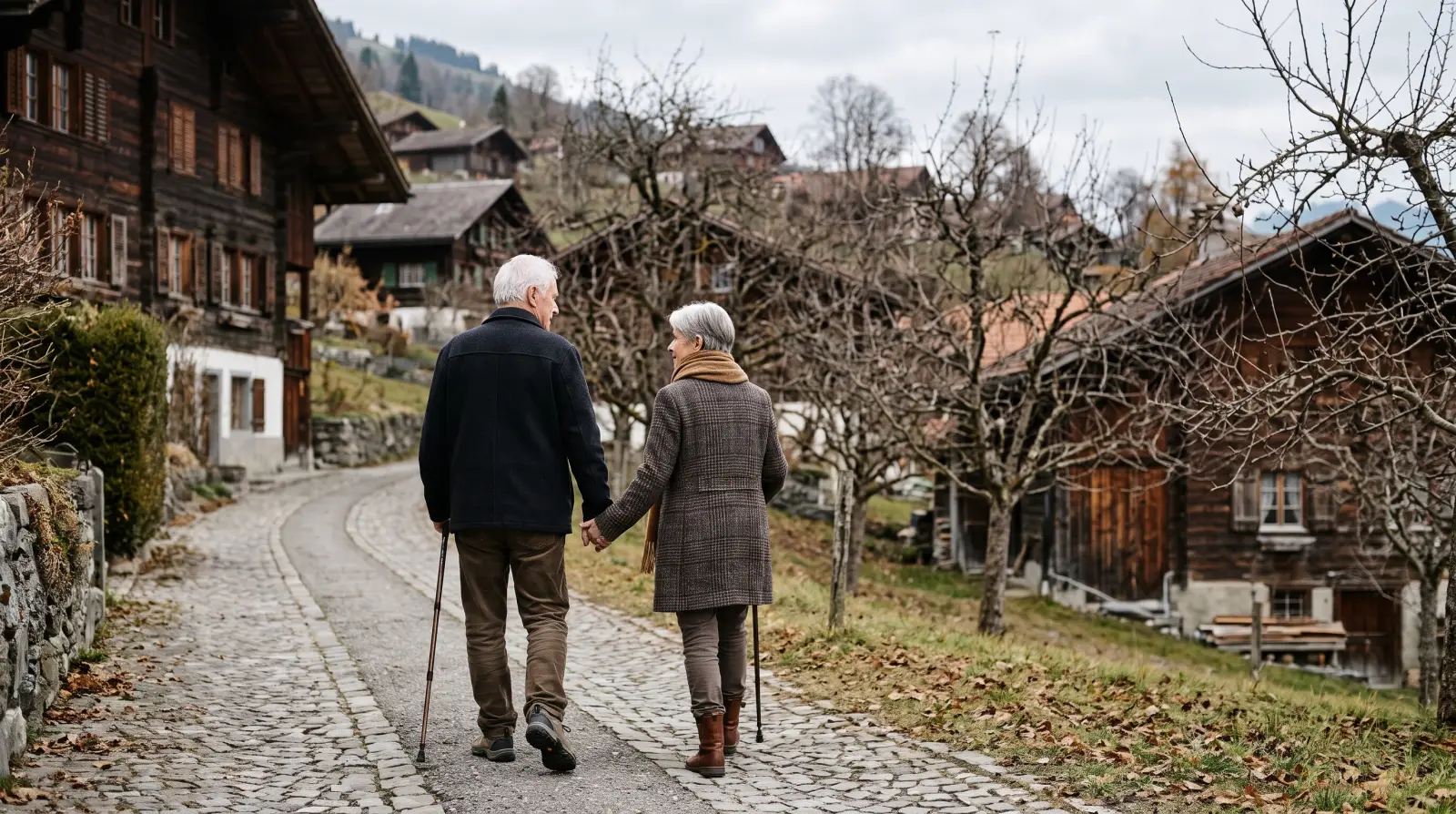 Ein älteres Paar spaziert Hand in Hand auf einem gepflasterten Dorfweg zwischen Holzhäusern und kahlen Obstbäumen.